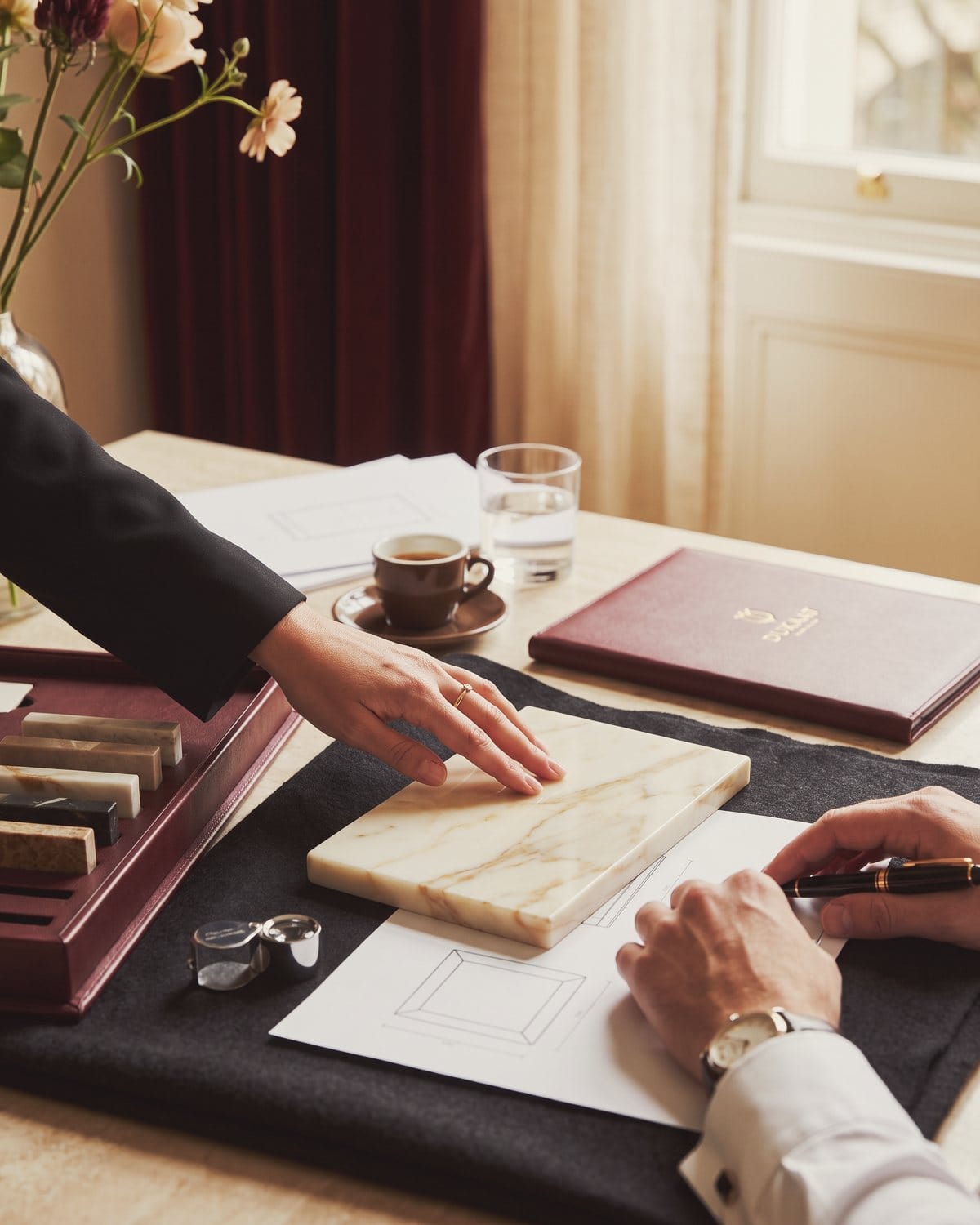 A hand runs over a polished marble sample beside a drawing and a loupe.
