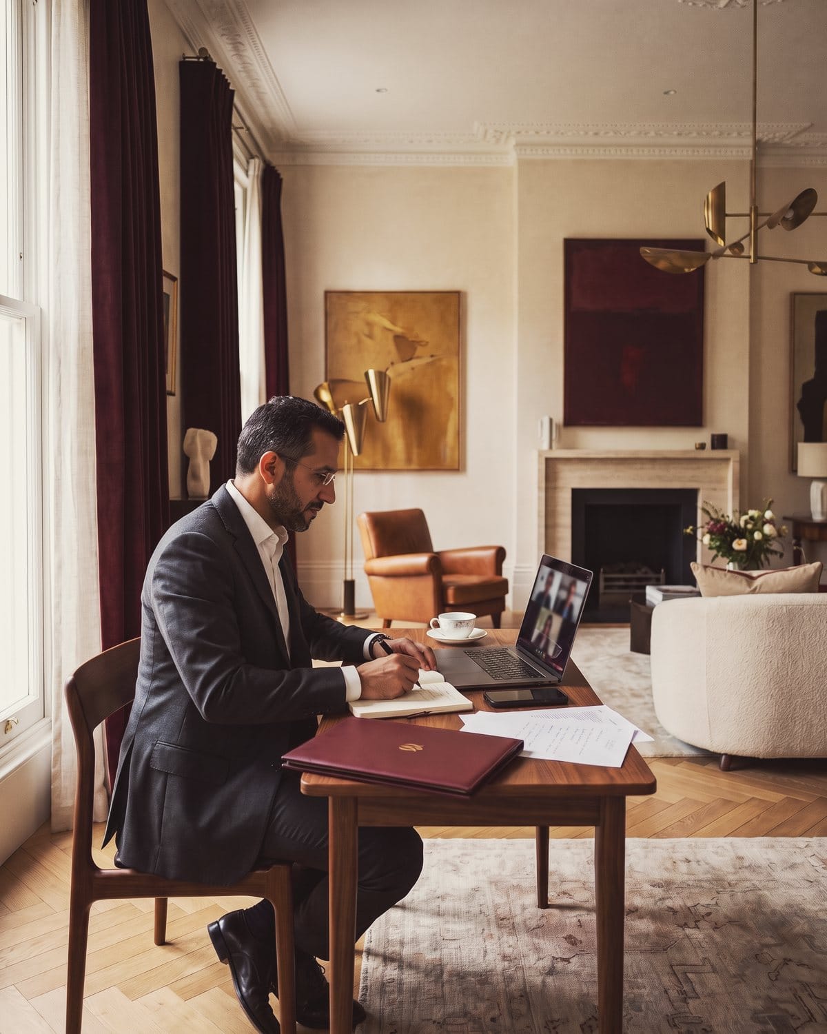 An adviser takes notes during a video call with partners abroad, at a table by the window of his study.