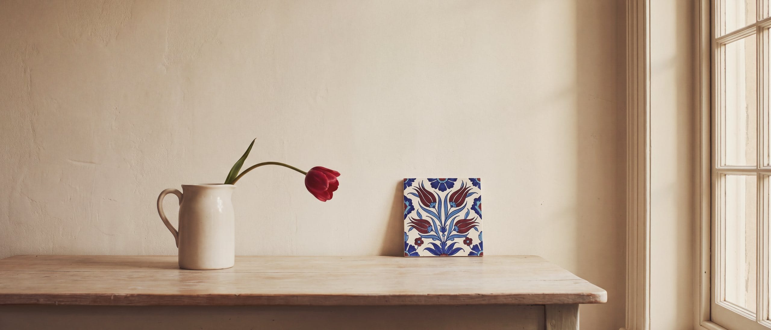 A single deep red tulip in a white earthenware jug beside a hand-painted tulip tile on a pale ash table.